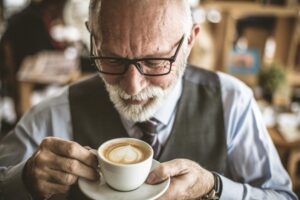 An older gentleman in a suit holding a cup of latte art coffee in a cozy café setting. Soft natural light fills the background.