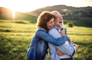 An older couple embraces tightly in a sunlit green field, capturing a moment of warmth and affection against a serene landscape.