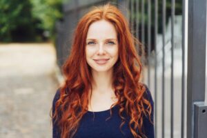 A woman with long, curly red hair stands in front of a blurred background with a metal gate, wearing a dark blue top.