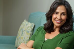 A woman with dark, wavy hair sits on a light blue couch, wearing a green sweater, with her arms crossed and a decorative pillow nearby.