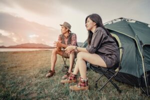 A man and woman sit on folding chairs near a green tent, enjoying nature by a lake at sunset, surrounded by grass and mountains.