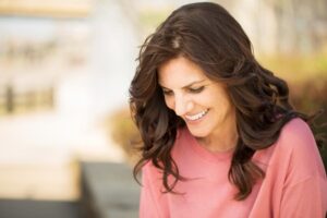 A woman with long, wavy brown hair wears a pink sweater, looking smile fully down in a softly blurred outdoor setting.