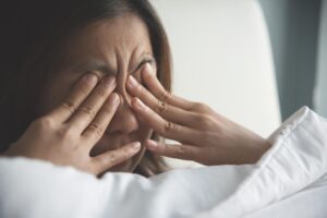 A woman rubbing her eyes while getting from sleep, still on her cozy white blanket on her bed .