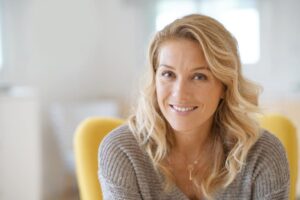 A woman with long, wavy blonde hair wears a cozy gray sweater, seated in a yellow chair.