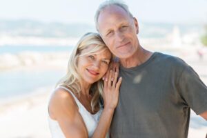 An elder couple stands close together on a sunny beach, smiling and holding hands.