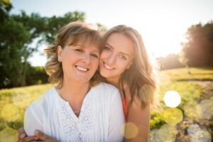 A woman in a white blouse embraces a young girl in a pink top, both smiling and surrounded by a sunlit, grassy landscape.