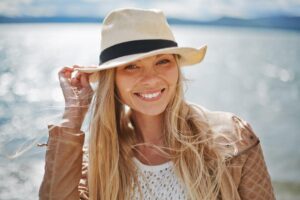 An woman wearing a straw hat and a tan jacket stands by a shimmering body of water, with gentle winds tousling their long hair.