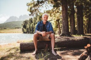 An older man sits on a log by a serene lake, surrounded by tall trees and mountains, enjoying a peaceful moment in nature.