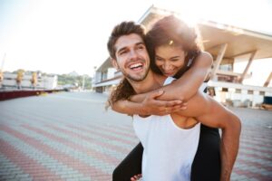 A joyful pair in a sunlit outdoor setting, with one person playfully giving a piggyback ride to the other, surrounded by modern architecture.