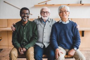 Three men sit closely together on a bench, each wearing casual attire, against a simple wooden background with minimal decor.