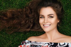A woman with long, wavy brown hair lies on green grass, wearing an off-shoulder patterned top, enjoying a serene moment outdoors.