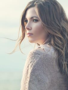 A woman with long, wavy hair stands outdoors, wearing a cozy, knitted sweater, with a soft, dreamy background of light and distant scenery.
