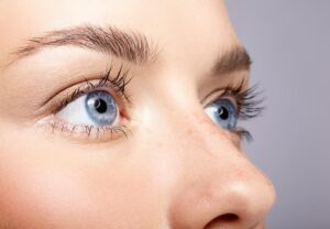 Close-up of a woman's eye with long eyelashes against a soft gray background, highlighting delicate skin tones and features.