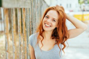 A young woman with wavy red hair poses outdoors against a weathered fence, wearing a casual gray top.