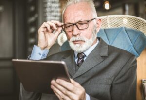 An older man in a suit sits in an elegant chair, holding a tablet and thoughtfully touching his glasses in a modern interior setting.