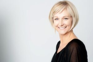 A woman with short blonde hair wearing a black top, posed against a light grey background, facing slightly to the left.