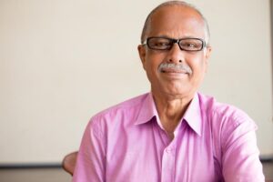 A senior man wearing a pink shirt sits in a well-lit room, exuding a calm and thoughtful presence against a neutral background.