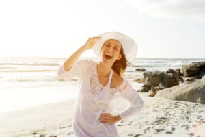 A woman in a white dress and sun hat poses on a sandy beach with waves crashing in the background under a bright sky.