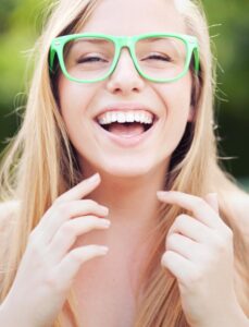 A young woman with long blonde hair wearing green sunglasses pointing her face with both hands, surrounded by a blurred natural background.