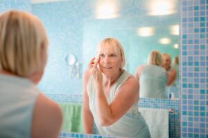 A woman with blonde hair is applying skincare in a bright blue-tiled bathroom, surrounded by mirrors and pastel towels.