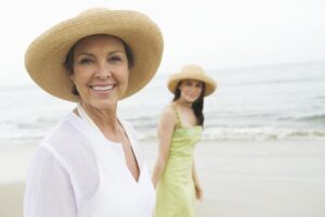 Two women stand on a beach, wearing sun hats. One in a white top, the other in a green dress, facing the ocean under a cloudy sky.