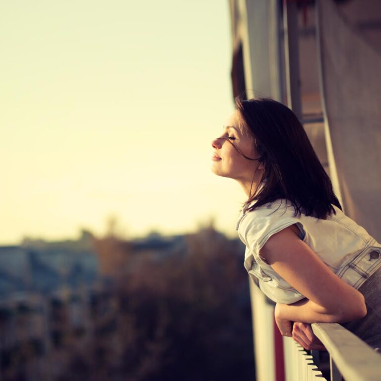 A young woman leans over a balcony, gazing thoughtfully into the horizon with a warm sunset in the background.