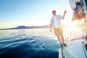 A person stands on the edge of a sailboat, gazing at a sunlit coastline with serene water and distant mountains under a clear sky.