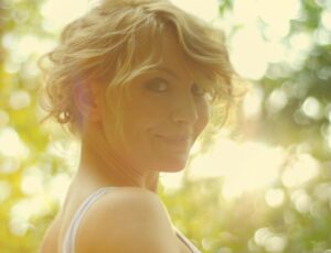 A woman with light, wavy hair stands with a serene expression in a sunlit, blurred natural background.
