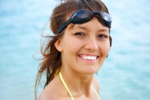 A woman with long, wavy hair stands by the water, wearing a yellow bikini top and a dark swimming goggles , set against a serene beach background.
