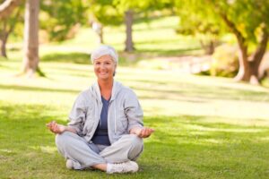 An older lady dressed in gray loungewear sits cross-legged on grass in a serene park, meditating with hands in a relaxed position.