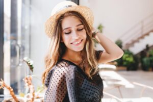 A woman with long, wavy hair wearing a straw hat poses stylishly in a bright, indoor cafe surrounded by greenery.