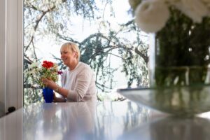 An older lady sits at a white table, arranging colorful flowers in a blue vase with a backdrop of greenery and sunlight streaming through a window.