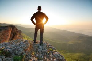 A person stands on a rocky cliff, gazing over a lush, mountainous landscape at sunrise, hands on hips, embracing the view.