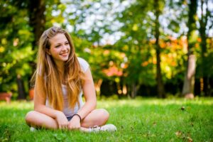 A girl sits cross-legged on green grass in a sunlit park, surrounded by trees with a blurred background of colorful leaves.