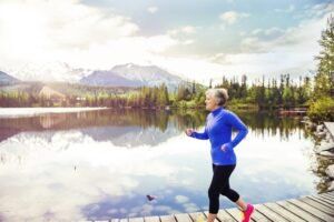 An older woman jogging along a wooden path by a serene lake, surrounded by mountains and lush green trees under a bright sky.