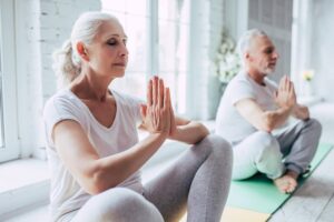 Two older adults practice yoga indoors, sitting cross-legged on mats with hands in a meditative gesture.