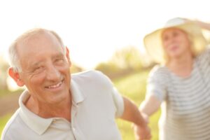 An elderly couple joyfully holds hands in a sunlit field, surrounded by greenery, sharing a moment of connection and happiness.