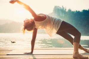 A woman practices yoga on a dock by a lake at sunset, stretching her body while the sun glimmers behind her.