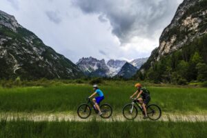 Two cyclists ride along a dirt path through lush green grass, surrounded by towering mountains and a cloudy sky.