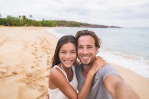 A couple takes a selfie on a sandy beach, with gentle waves lapping at the shore and lush greenery in the background.