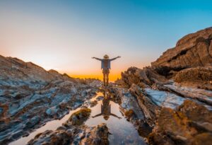 A person in a hat stands on rocky terrain, arms outstretched, reflecting in shallow water as the sunset casts a warm glow.