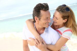 A couple embraces playfully on a beach, with the ocean and sky in the background, exuding joy and affection.