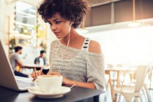 A woman wearing a cozy, off-shoulder sweater is focused on their laptop, with a cup of coffee beside them in a bright cafe.