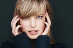 A girl with long, light brown hair holds her head, wearing a dark sweater, against a blurred background.