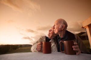 Two older couple sit together, holding wooden mugs, against a beautiful sunset backdrop with soft clouds and rolling hills.