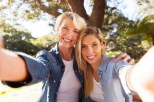 Two women take a cheerful selfie in a sunlit park, surrounded by trees, wearing casual outfits and smiling together.