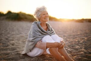 An elderly woman with curly hair sits on the beach, wrapped in a cozy shawl, as the sun sets in the background, casting a warm glow.