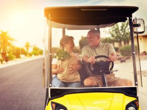 A couple enjoys a sunny ride in a yellow golf cart, with a paper bag of fresh greens on their lap, surrounded by a tranquil neighborhood.